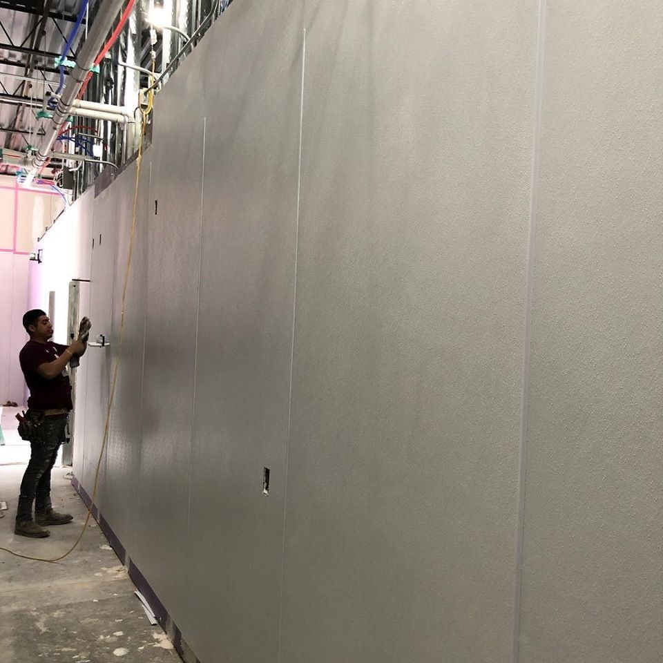 Worker installing sections of a gray wall with visible wiring and pipes above in a partially constructed building.